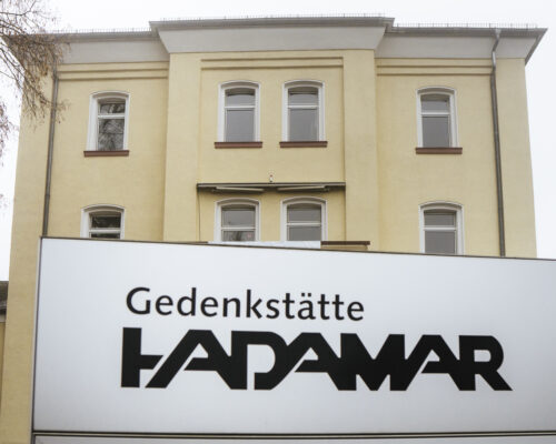 Ein Schild mit der Aufschrift "Gedenkstätte Hadamar". Im Hintergrund ein großes gelbliches Gebäude - die Gedenkstätte.