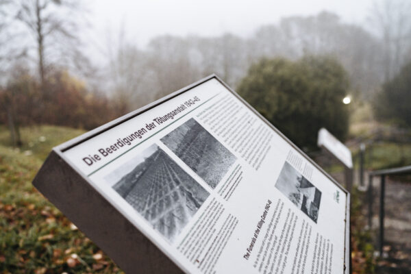An outdoor exhibition panel entitled "The funerals at the killing centre 1942-1945". Another panel can be seen in the background although it is blurred.