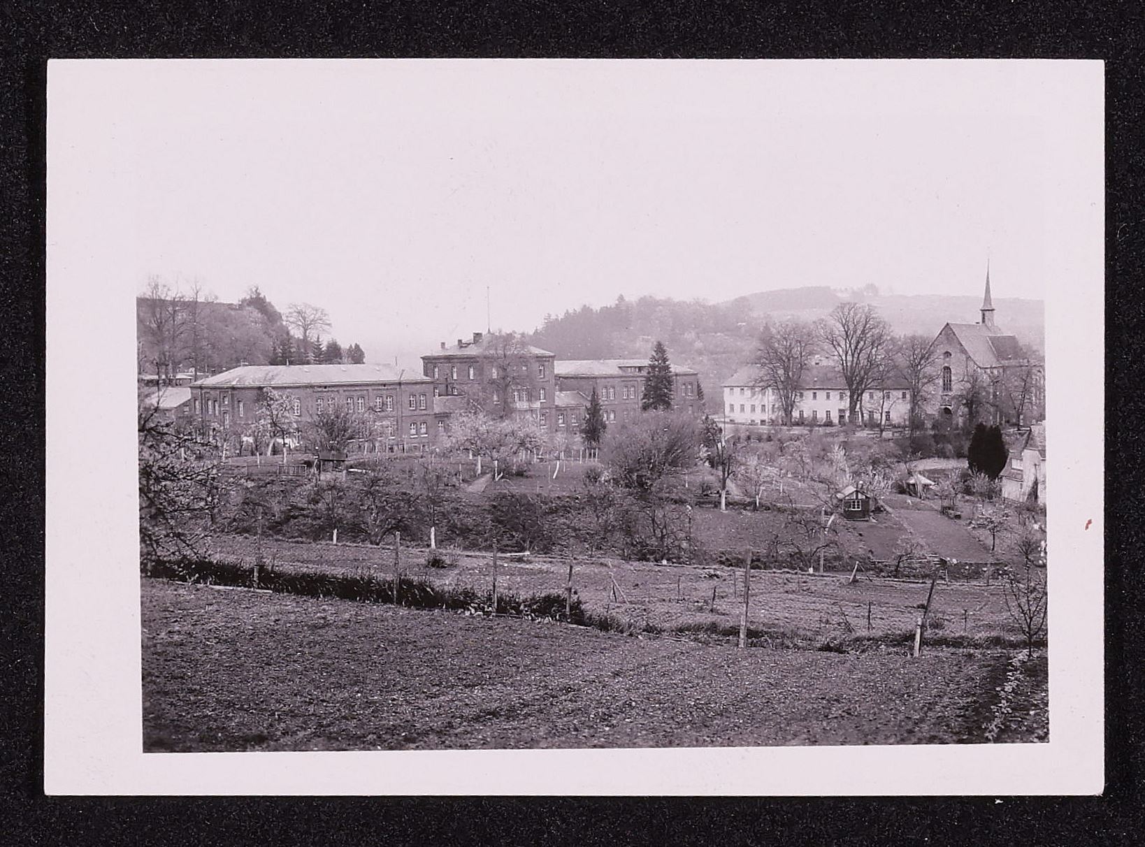 Black and white photo. A field is in the foreground of the photo; a large building as well as a monastery is in the background.