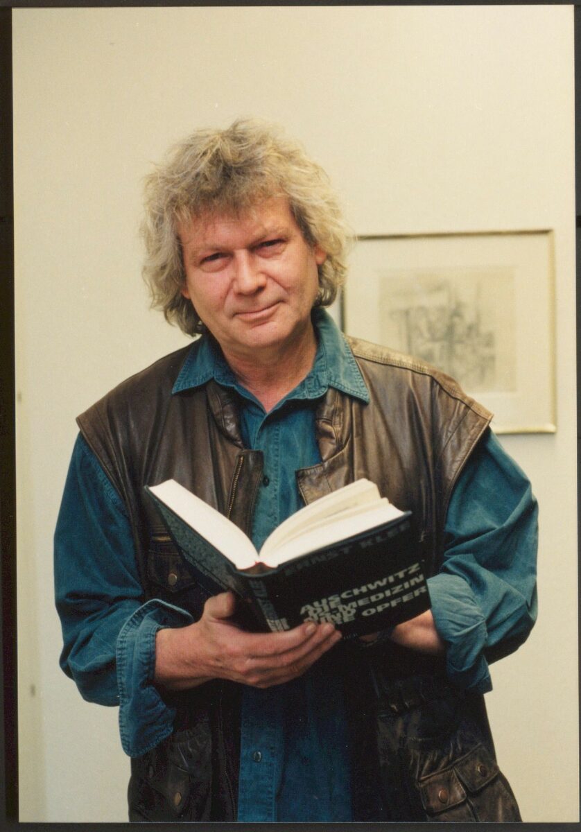 Farbfoto eines älteren Mannes mit mittellangen grauen Haarzen der ein Buch in den Händen hält. Vom Titel ist nur das Wort "Auschwitz" zu lesen. Portrait von Ernst Klee.
