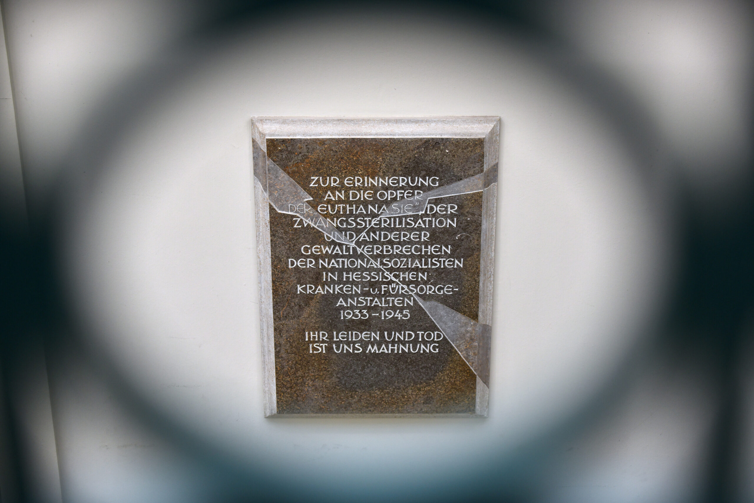 Colour photograph of a brown plaque embedded in a wall. The effect that is created is as if the plaque were made of glass and has a large crack running through it. Translated, the inscription reads "In remembrance of the victims of "euthanasia", forced sterilization and other violent crimes committed by the Nazis in Hessian hospital and care facilities from 1933-1945. Their suffering and death is a warning to us all."