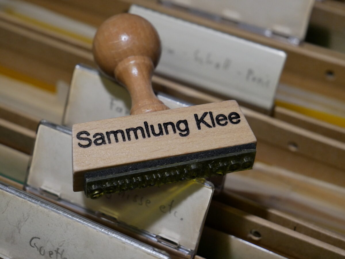 Farbfoto eines hölzernen Stempels mit der Aufschrift "Sammlung Klee". Im Hintergrund unscharf Akten.