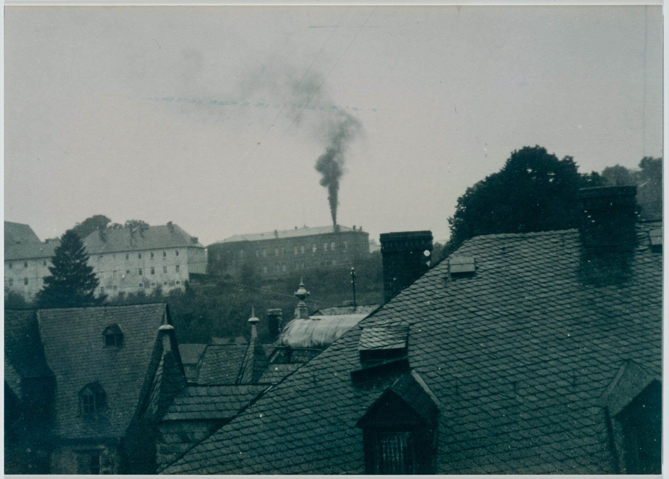 Black and white photo of the Hadamar institution from afar. In the foreground are the roofs of several buildings in Hadamar. A black smoke column is rising into the sky from the chimney of the institution.