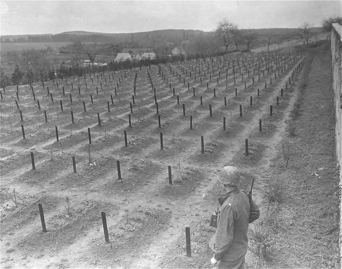 Schwarz-Weiß-Foto. Ein US-amerikanischer Soldat steht am rechten Bildrand mit dem Rücken zur Kamera und blickt auf einen Friedhof mit mehreren Reihen schmuckloser Einzelgräber.