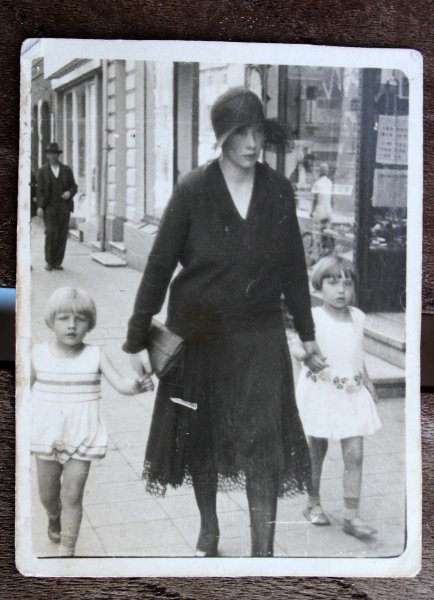 Eine schwarz-weiß Fotografie einer Frau und zwei Kindern. Die Frau trägt ein schwarzes Kleid und einen schwrazen Hut. An der Hand hält sie zwei Kinder, die weiße Sommerkleider tragen. Das Bild wurde auf der Straße aufgenommen, man sieht eine Hauswand und ein Ladengeschäft im Hintergrund. Die Personen schauen nicht in die Kamera, sondern rechts daran vorbei.