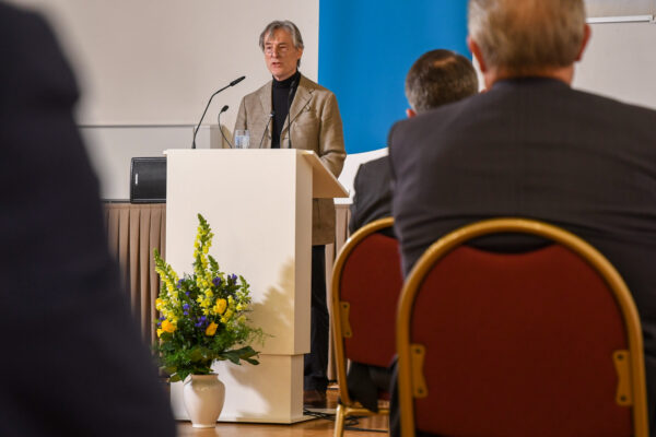 Ein Mann steht an einem Rednerpult und spricht, das Foto wurde aus dem Publikum aufgenommen.