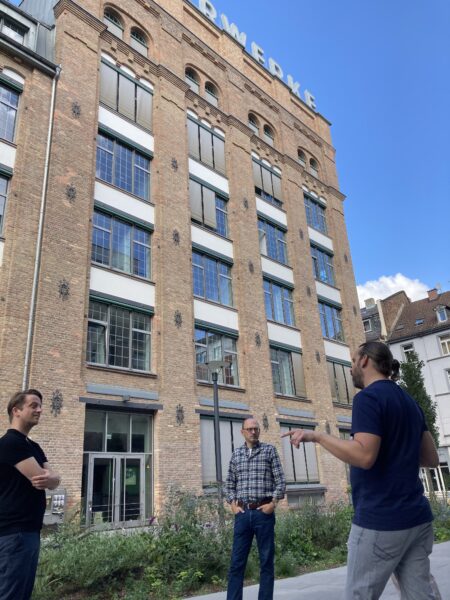 Eine Gruppe von drei Männern steht vor einem großen Backsteingebäude. Sie stehen in einem Kreis und sprechen miteinander, hinter ihnen ragt das Gebäude hoch.