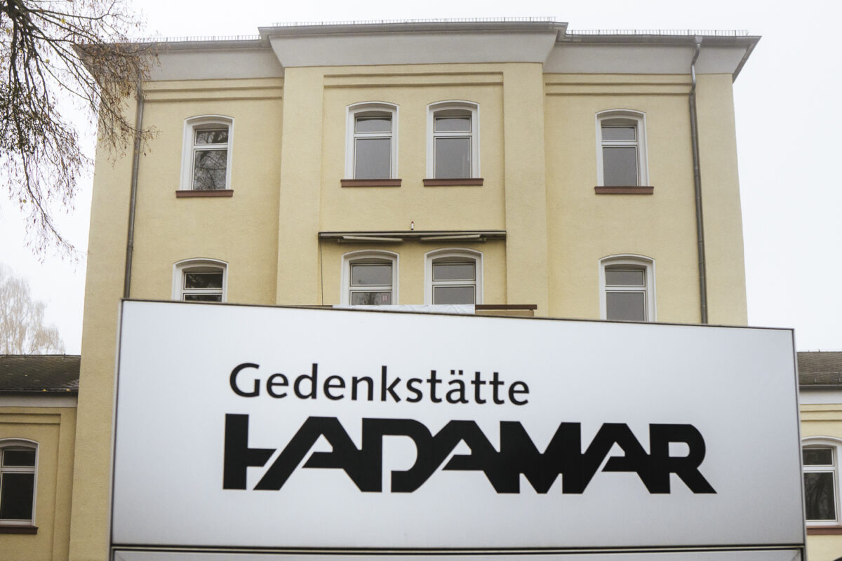 Ein Schild mit der Aufschrift "Gedenkstätte Hadamar". Im Hintergrund ein großes gelbliches Gebäude - die Gedenkstätte.