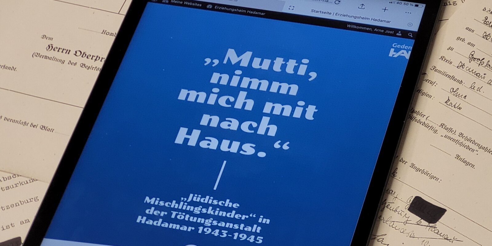 Ein Tablet liegt auf Akten. Auf dem Tablet ist eine Website geöffnet, man kann den Titel "Mutti nimm mich mit nach Haus" in weißer Schrift auf blauem Hintergrund lesen.