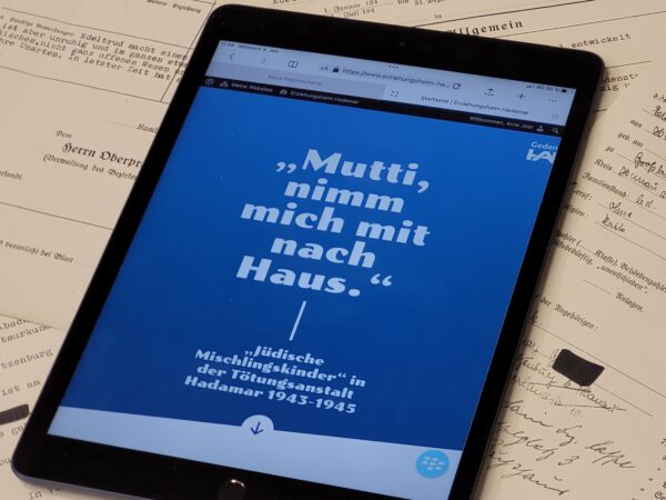 Ein Tablet liegt auf Akten. Auf dem Tablet ist eine Website geöffnet, man kann den Titel "Mutti nimm mich mit nach Haus" in weißer Schrift auf blauem Hintergrund lesen.
