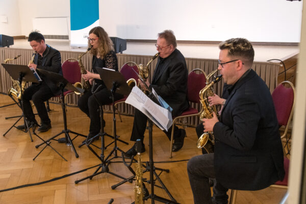 Ein Ensemble bestehend aus fünf Personen, die das Saxophon spielen. Vor ihnen stehen Notenständer, sie sitzen vor eine Bühne.