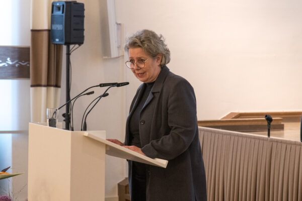 Eine Frau mit grauen Haaren und einem dunklen Hosenanzug steht vor einem Rednerpult und schaut in die linke Ecke des Bildes.