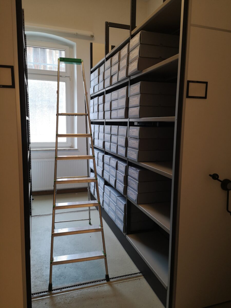 Ein Foto der Archi-Außenstelle des LWV-Archivs. In beweglichen Regalen stehen graue Kartons, in denen sich die Akten befinden. Auf den Kartons sind Schilder befestigt, auf denen die enthaltenen Aktennummern zu lesen sind. Zwischen den Regalen steht eine Leiter.