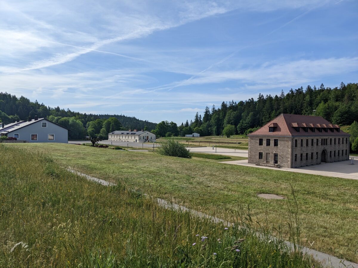 Ein Foto eines alten Gebäudes, das von Wiesen umgeben ist. Auf der linken Seite des Bildes ist ein Wohnhaus zu sehen.
