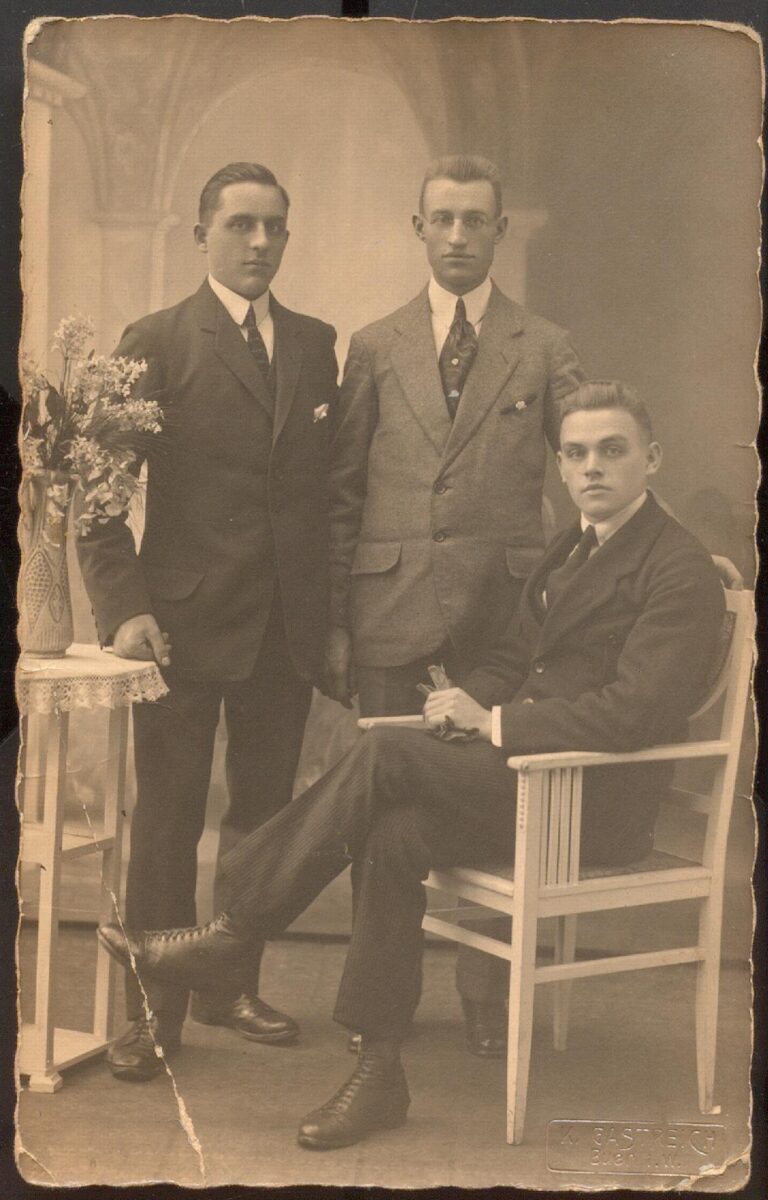 Ein schwarz-weiß Foto von drei Männern. Zwei von ihnen (der linke und der Mann in der Mitte) stehen, der rechte Mann sitzt auf einem weißen Stuhl. Neben dem linken Mann ist ein weißer Tisch zu sehen, auf dem Blumen in einer Vase stehen. Die drei Männer haben Anzüge an und schauen in die Kamera.