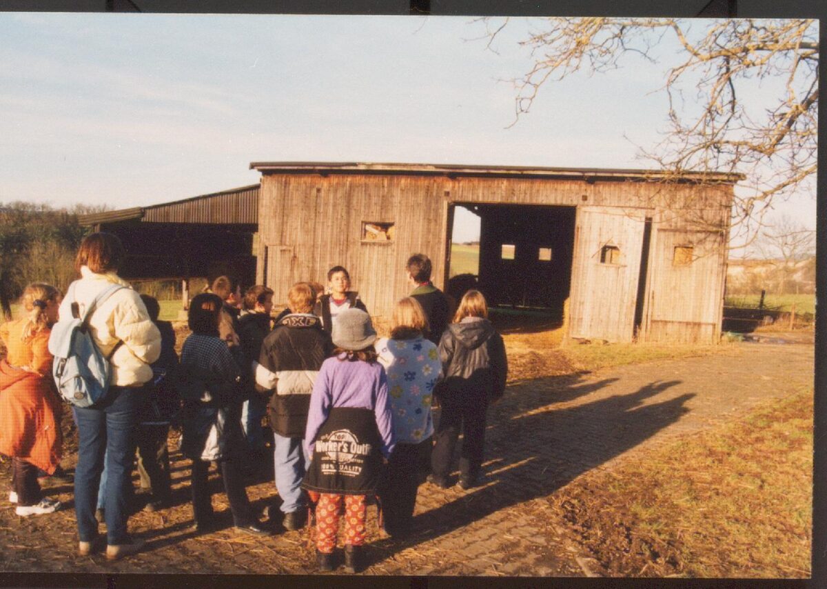 Eine Gruppe Schülerinnen und Schüler steht vor der ehemaligen Busgarage auf dem Feld. Die Garage hat das mittlere Tör geöffnet, auf der Rückseite ist auch ein Tor offen. Neben der Garage ist ein hölzerner Unterstand angebaut.