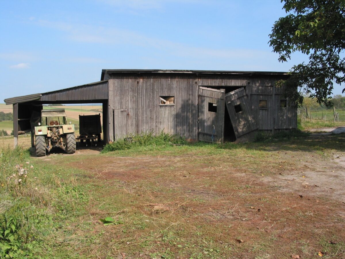 Die hölzerne Busgarage auf einem Feld. Das mittlere Tor hängt schief in den Angeln, links neben der Garage ist ein Unterstand angebaut, unter dem ein Traktor steht.