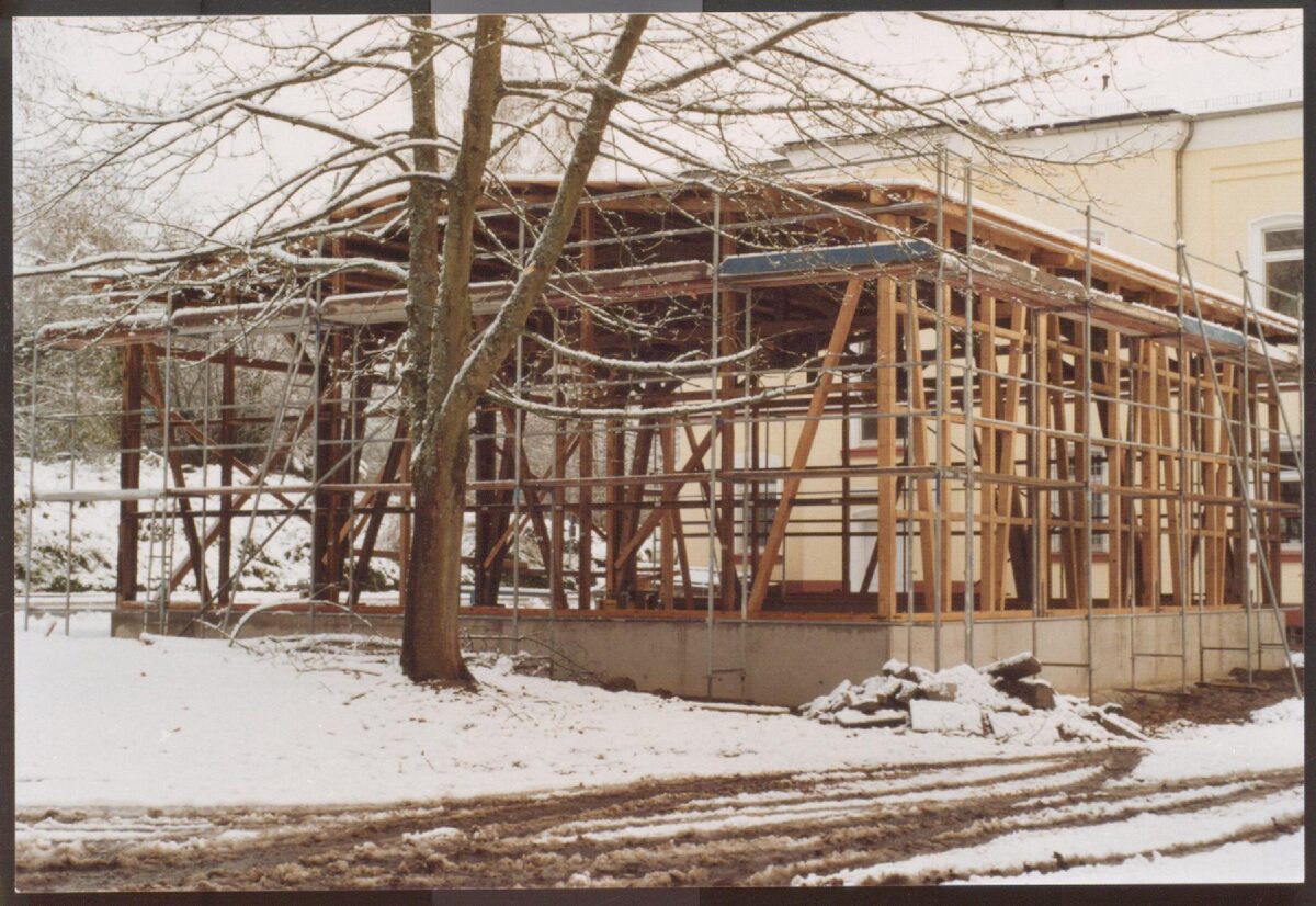 Ein Foto der ehemaligen Busgarage beim Wiederaufbau im Innenhof der Gedenkstätte. Es liegt Schnee. Die Garage ist noch nicht vollständig aufgebaut, bisher steht lediglich das Grundgerüst und das Dach.