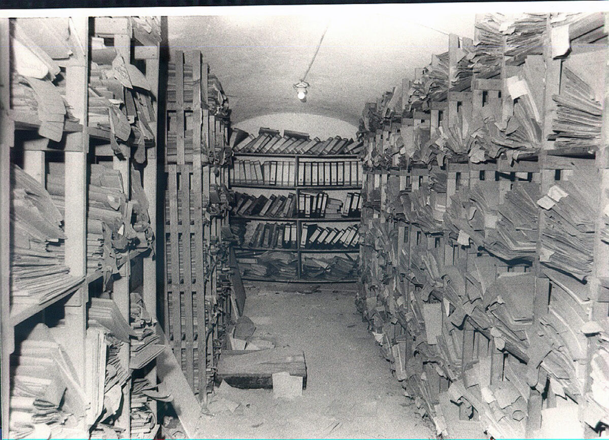 Schwarz-weiß Foto von einem Kellerraum. An den Wänden stehen Regale, in denen bis zur Decke Patientenakten gestapelt sind.