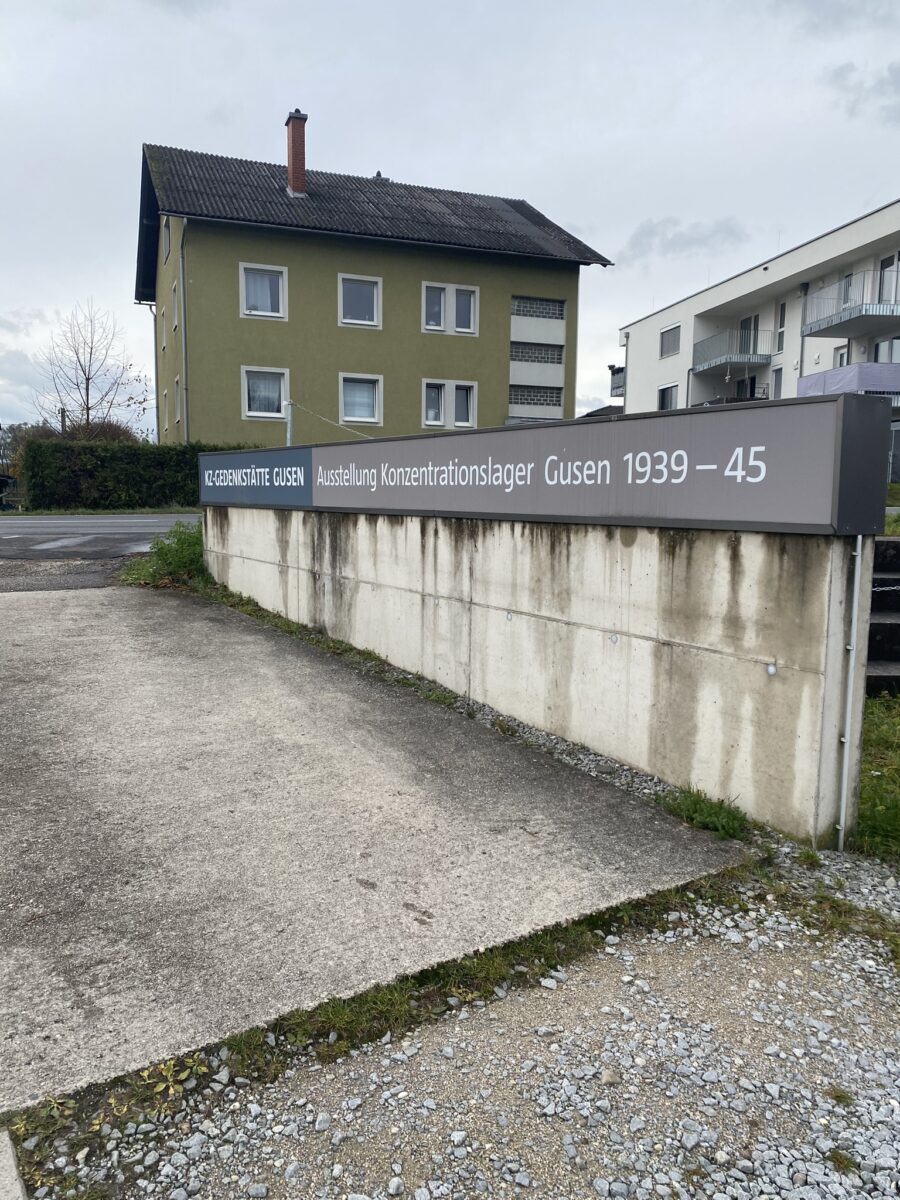 Der Eingang zur Gedenkstätte Gusen ist zu sehen. Neben der Einfahrt in ein großes Schild mit der Inschrift „KZ-GEDENKSTÄTTE GUSEN Ausstellung Konzentrationslager Gusen 1939-45“ befestigt. Im Hintergrund sind die Hauptstraße und Wohngebäude zu sehen.
