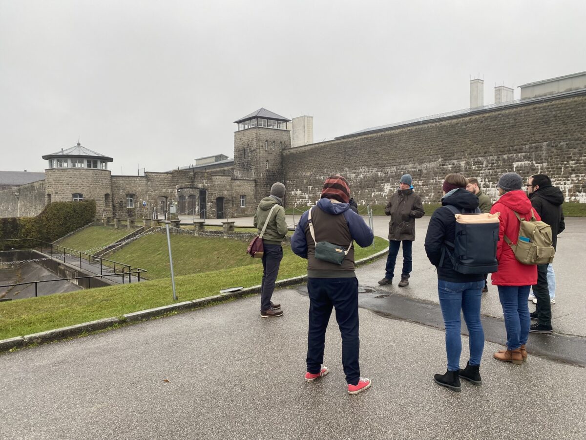 Eine Gruppe von Menschen steht im Kreis auf einem asphaltierten Platz vor einem Gebäude, das wie eine Festung aussieht. Die Mauern und Wachtürme sind aus Stein gebaut. Dies ist das ehemalige Konzentrationslager Mauthausen. Die Gruppe schaut zu einem Mann, der spricht und sich gerade wegdreht um auf eine Stelle hinter sich zu schauen.