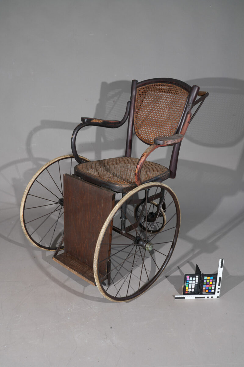 Ein historischer Rollstuhl aus Holz, der vor einem grauen Hintergrund steht. Neben dem Rollstuhl auf dem Bodem liegt eine Farbpalette.
