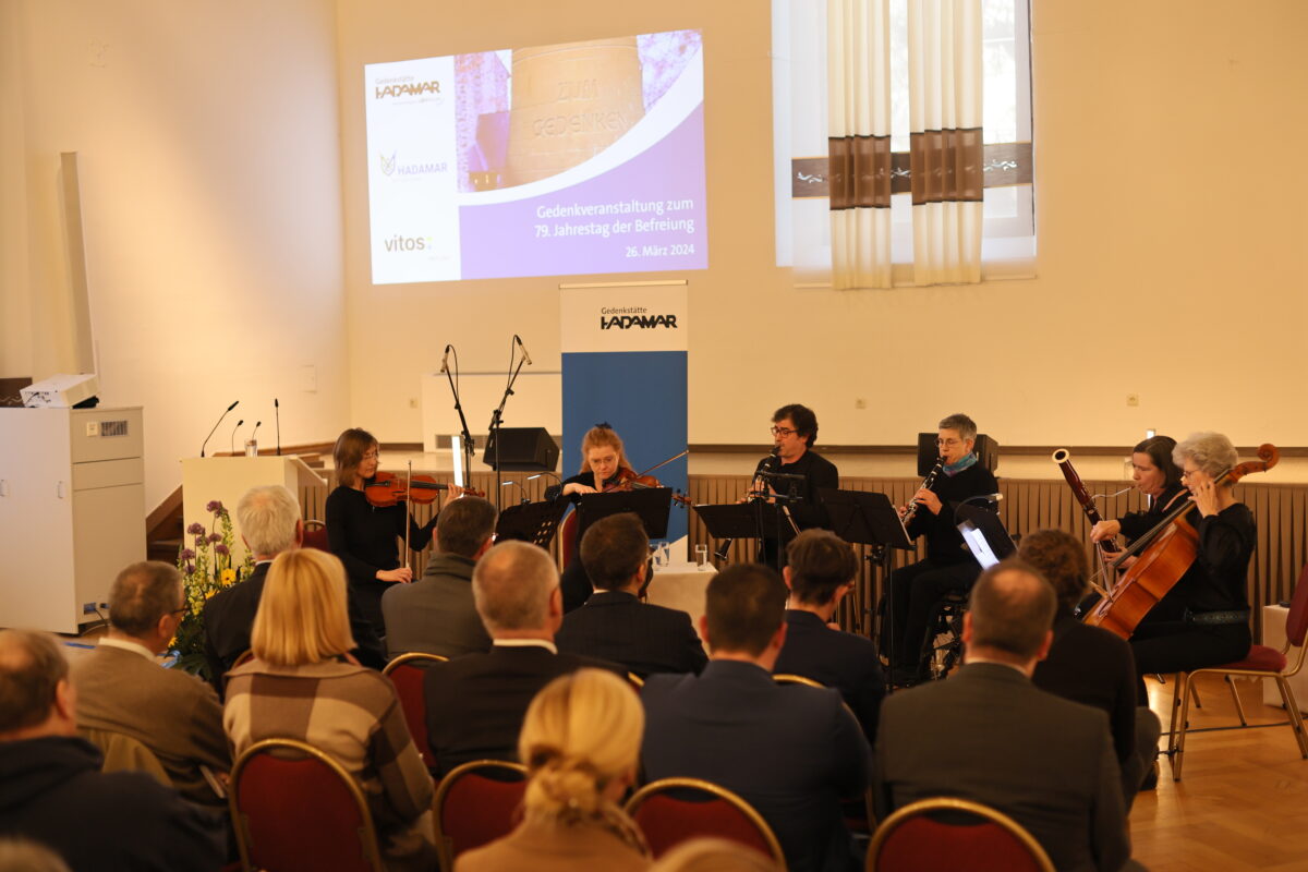 Ein Foto von einer Veranstaltung. Vor einer Bühne sitzen sechs Musikerinnen und Musiker mit verschiedenen Instrumenten. Davor sind Menschen im Publikum zu sehen.